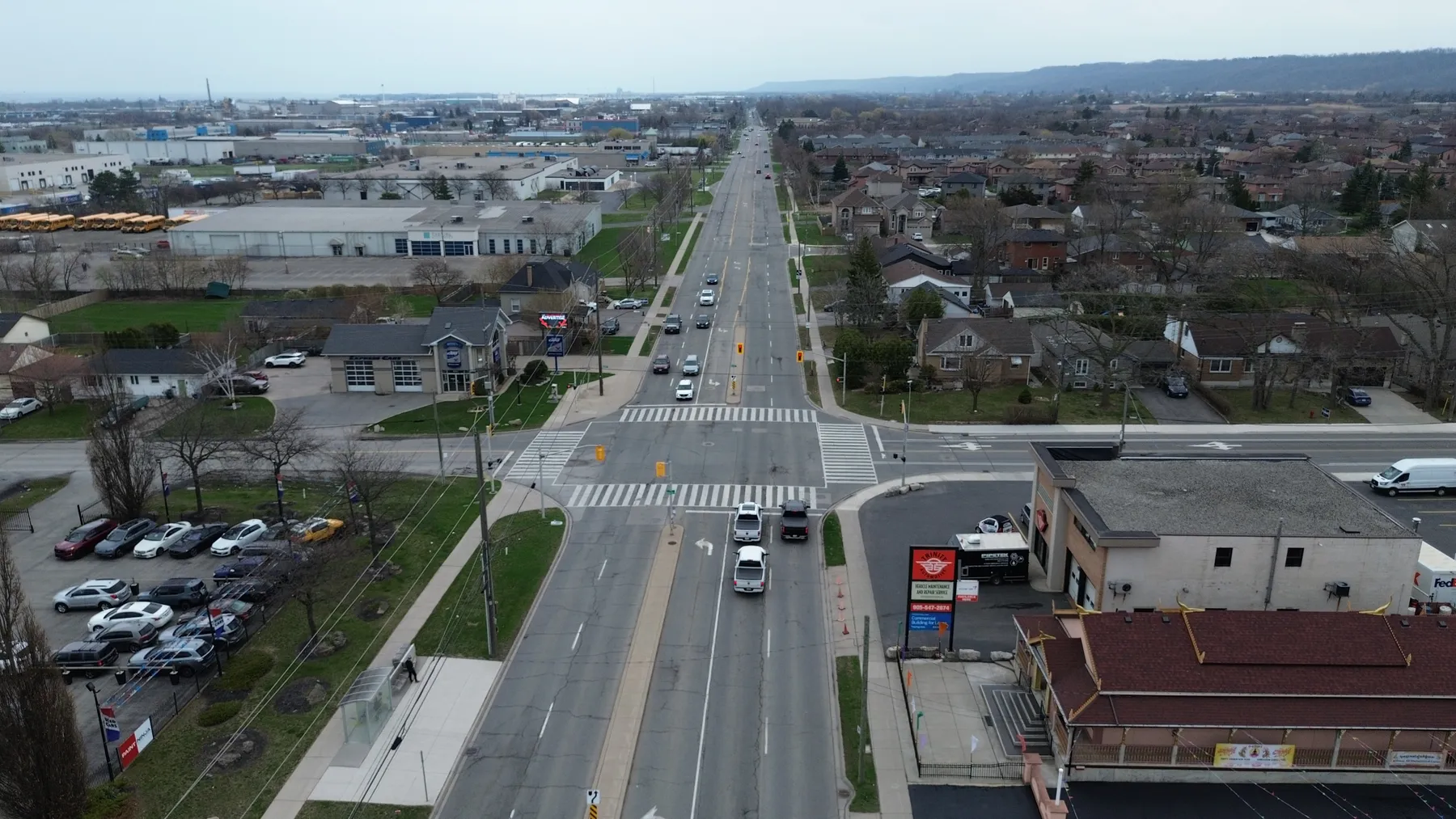 Low aerial along Barton Street East toward the junction, with the 521 Barton digital billboard over the eastbound lanes and the corner property on the right