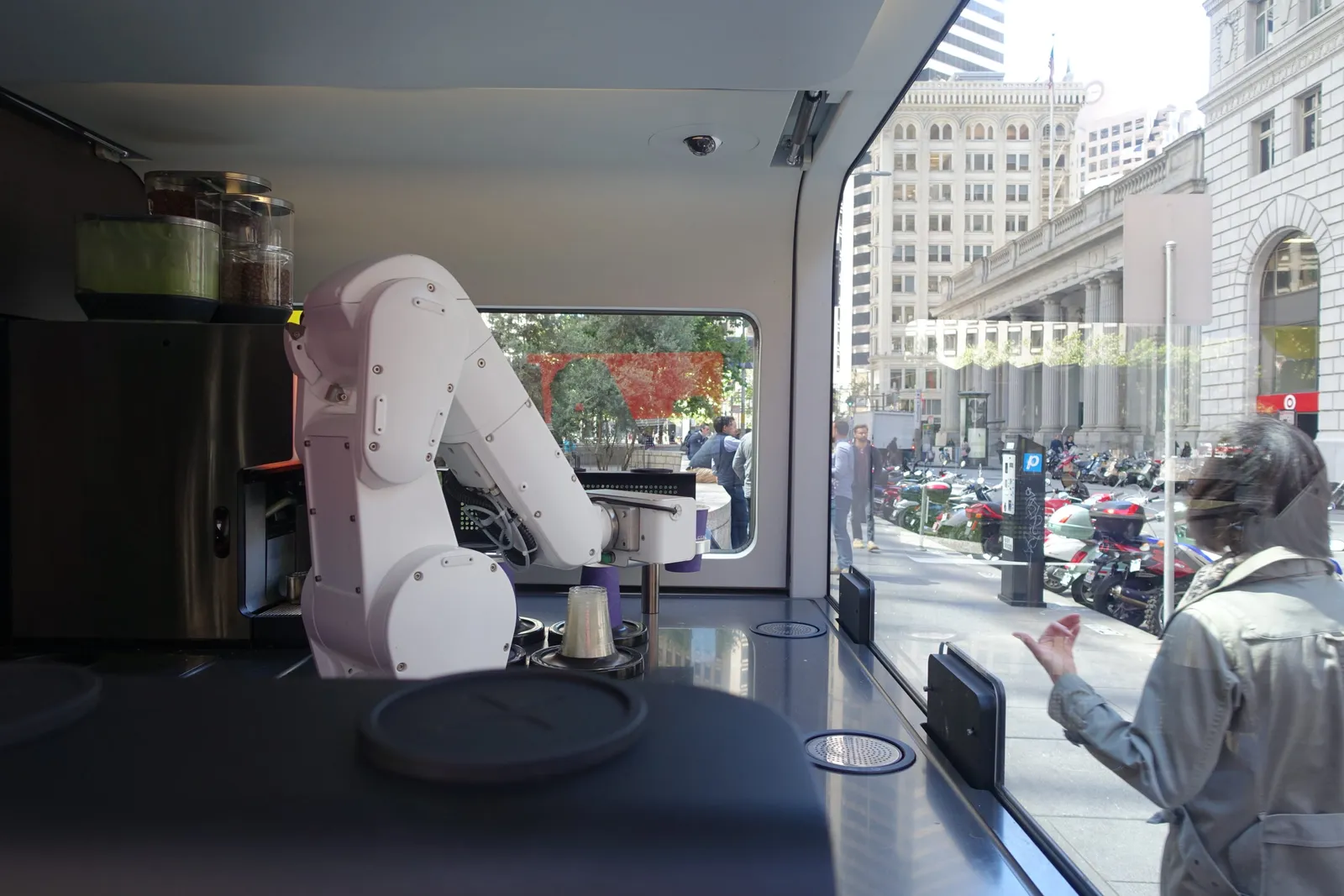 Inside the Cafe X kiosk: the white six-axis robot arm at the espresso modules, a customer watching through the glass