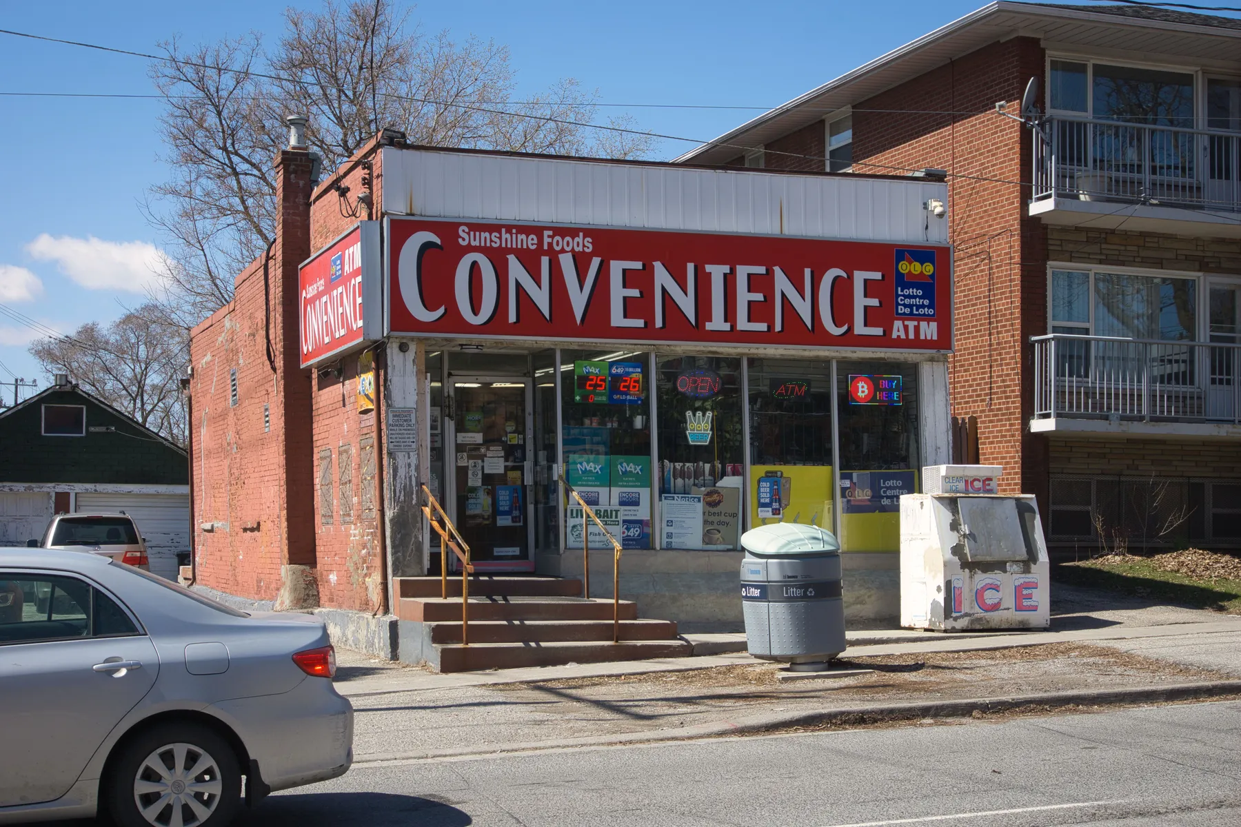 An independent corner convenience store in Toronto with a hand-painted sign, lotto decals, and an ice freezer out front