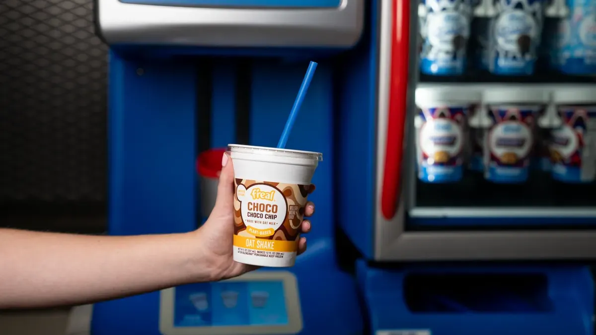 A hand holds a pre-portioned frozen shake cup in front of an in-cup blending machine and cup freezer
