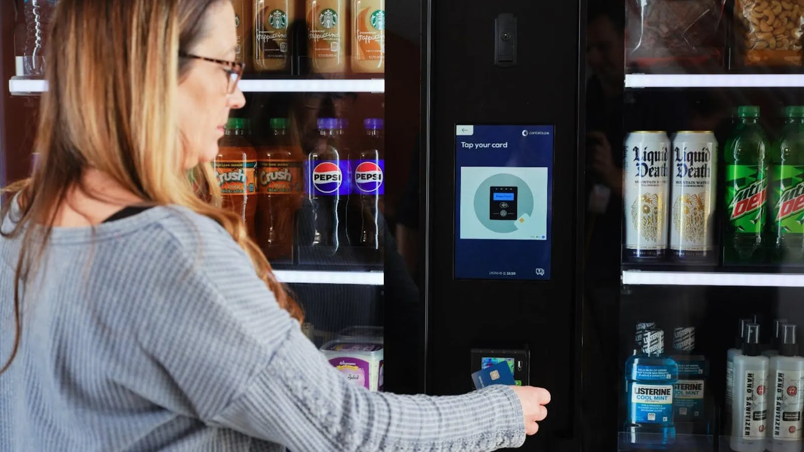A customer taps a payment card to unlock a smart cooler stocked with prepackaged drinks and snacks