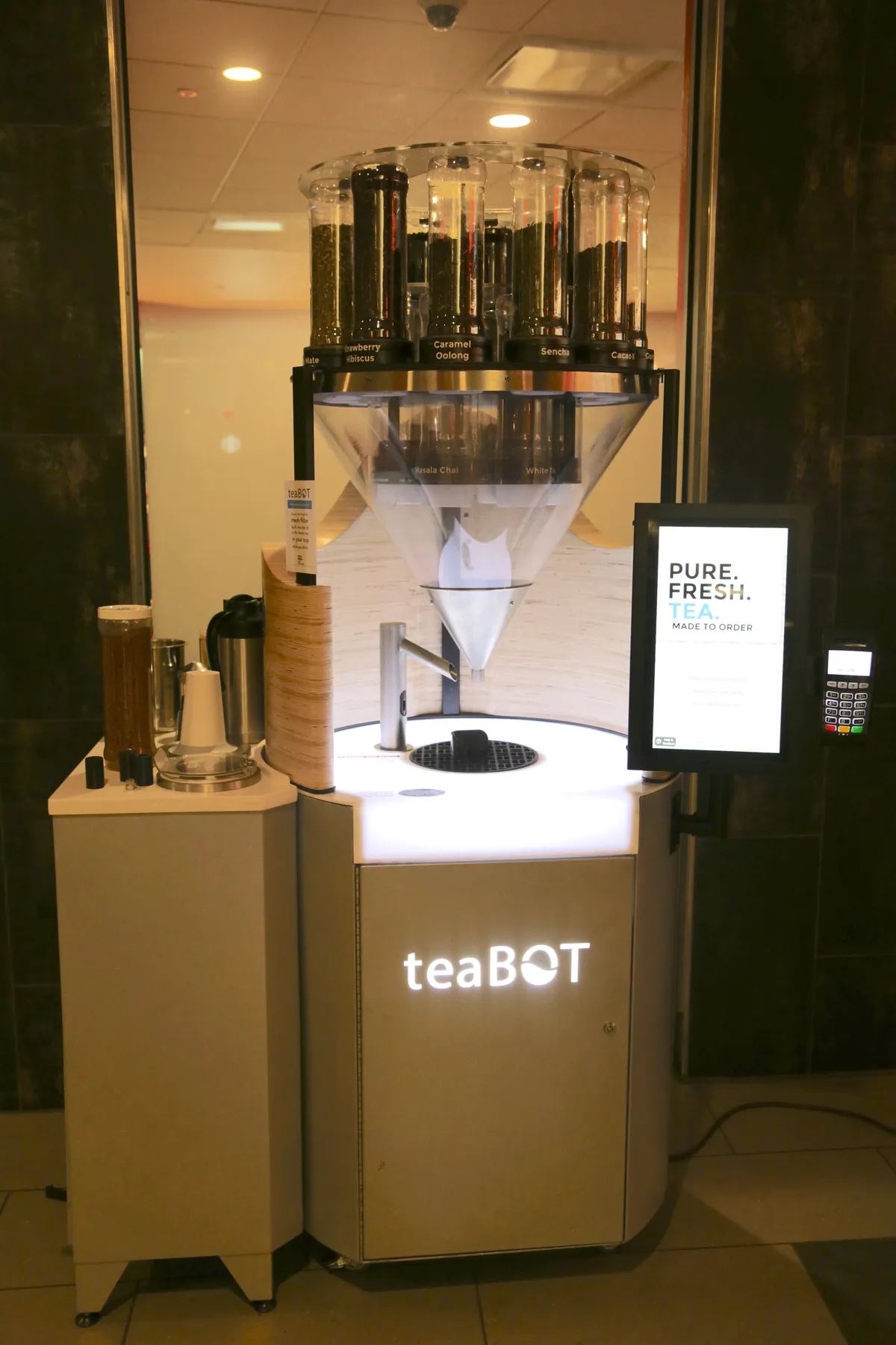 A teaBOT automated loose-leaf tea kiosk with clear tea canisters above a lit dispensing counter