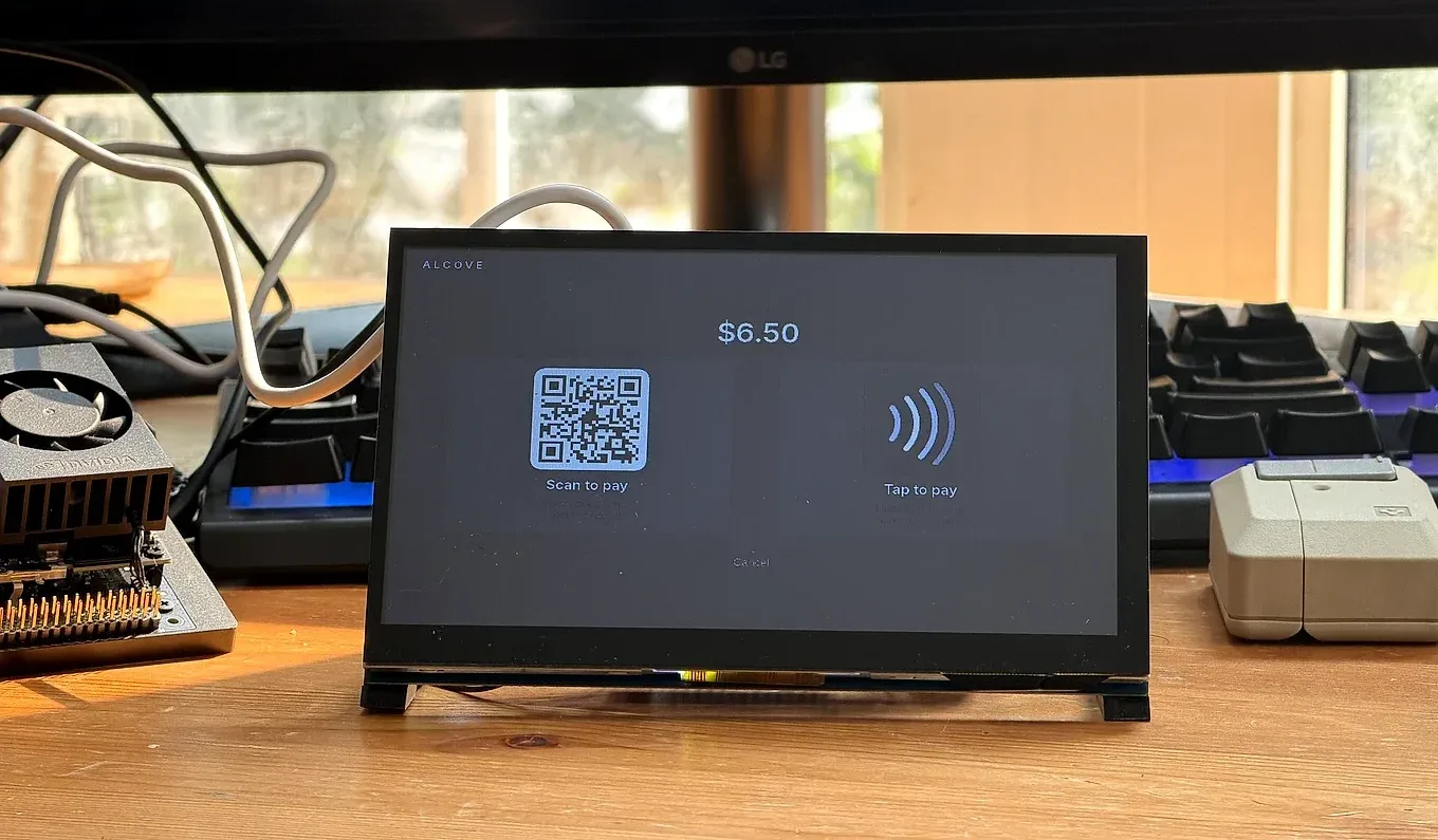The Alcove bench unit on a workbench, its touchscreen showing checkout: total $6.50 with a scan-to-pay QR beside a contactless tap-to-pay symbol.